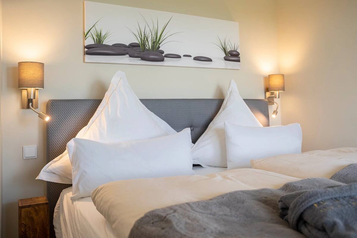 A hotel room with a bed, two wall-mounted lamps, a headboard, and a painting of rocks and plants.