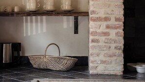 Een keuken met een bakstenen open haard, een plank met kopjes en schotels, een mand en een roestvrijstalen waterkoker.