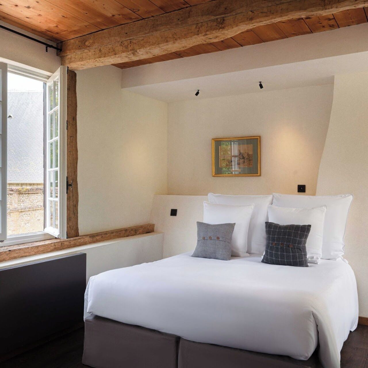 A bedroom with a wooden ceiling, a bed with white bedding, a window with a view, and a framed picture on the wall.