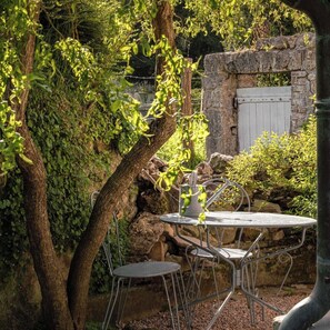 Un espacio de descanso al aire libre de estilo rústico, con pared de piedra, mesa y sillas de metal, y un árbol con follaje verde.