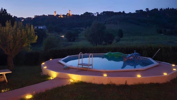 Une piscine avec un escalier, entourée d’un petit muret illuminé, située dans un espace verdoyant agrémenté d’arbres et avec une colline en arrière-plan.