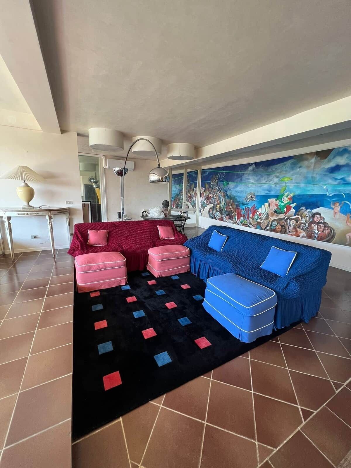 Ein Wohnzimmer mit einem roten und blauen Sofa, einem schwarzen Teppich mit geometrischen Mustern und einem großen Wandgemälde, das eine Menschenmenge zeigt.