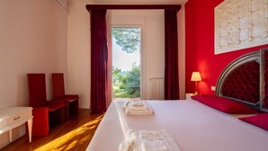 Quarto de hotel com cama, escrivaninha, cadeira, janela com cortinas e luminária.
