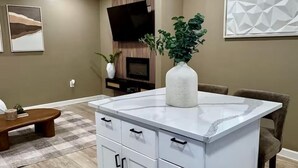 A modern kitchen with white cabinetry, a central island, and a flat-screen TV mounted on the wall. - Walk to DPAC + Bulls - Chic Downtown Durham condo (Durham)