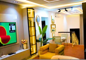 Een moderne woonkamer met een flatscreen-tv, een gele bank, een plant en een eethoek met een tafel en stoelen.