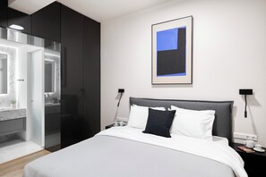 A modern hotel room with a bed, bedside tables, a nightstand, a framed picture, and a bathroom visible through a glass door. - Brand New Modern Apartment- Spacious Living & 2 Bathrooms (Glifada)