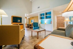 A living room with a sofa, armchair, coffee table, TV, and a bookshelf. - Bright 2-bedroom apartment with AC in lovely Palm Desert Breezes (Palm Desert)