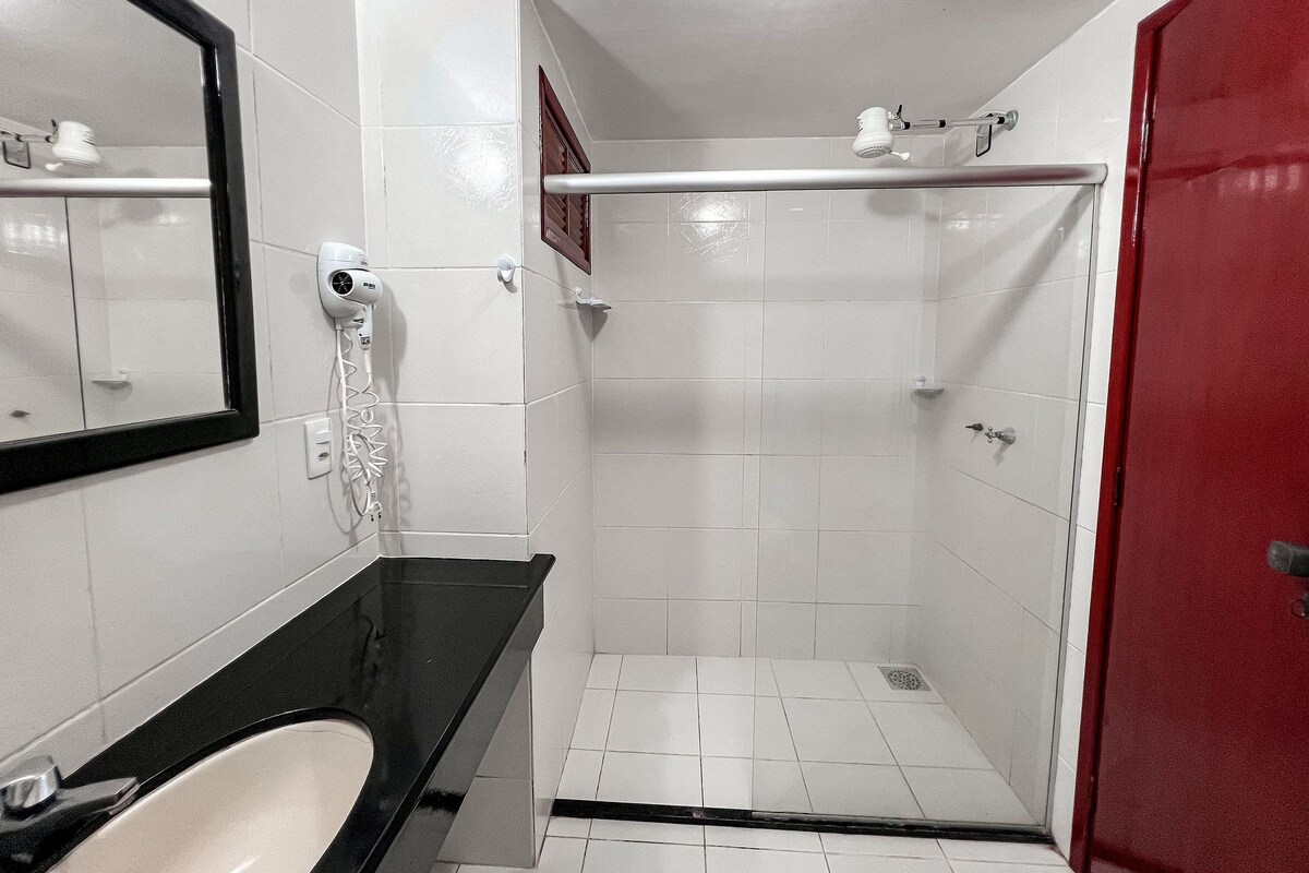 Un baño con un lavamanos blanco, un espejo, una ducha con puerta blanca y una puerta roja.