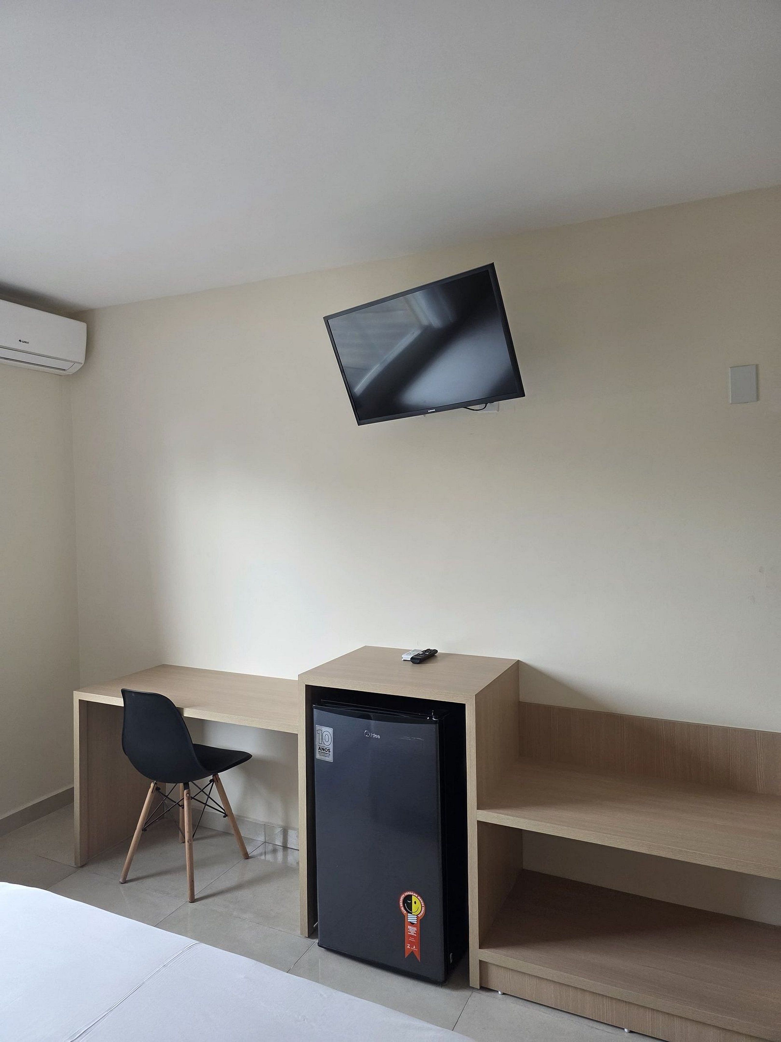 A compact room with a bed, a desk with a chair, a small black refrigerator, and a mounted TV.