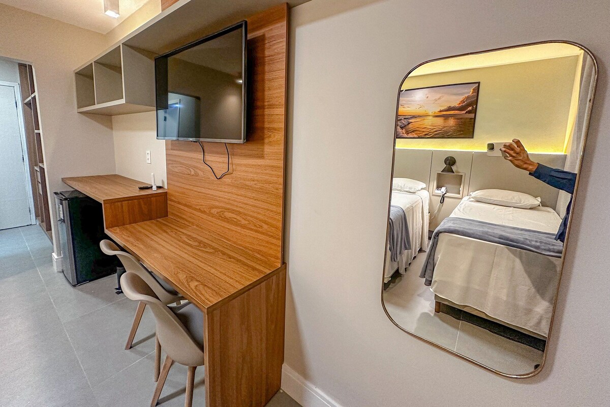 A hotel room with a wooden desk, a flat-screen TV, a bed with a white headboard, and a large mirror reflecting the room.