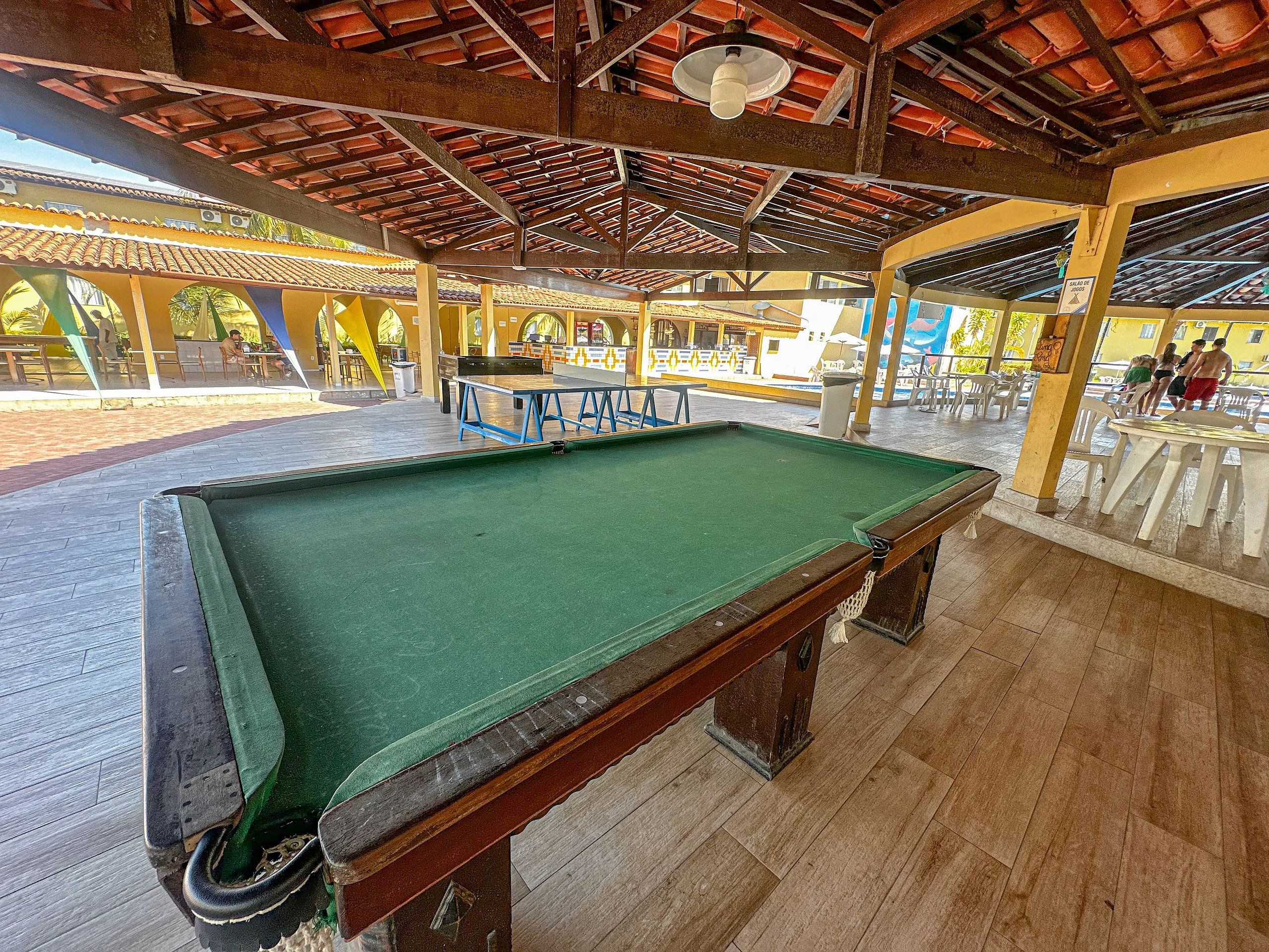 A pool table in a covered area with a wooden floor and a ceiling with a red-tiled roof.