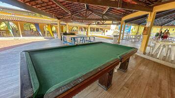 A pool table in a covered area with a wooden floor and a ceiling with a red-tiled roof.