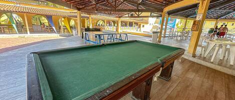 A pool table in a covered area with a wooden floor and a ceiling with a red-tiled roof.