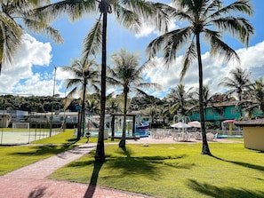 Um resort tropical com palmeiras, área esportiva cercada e piscina com guarda-sóis.