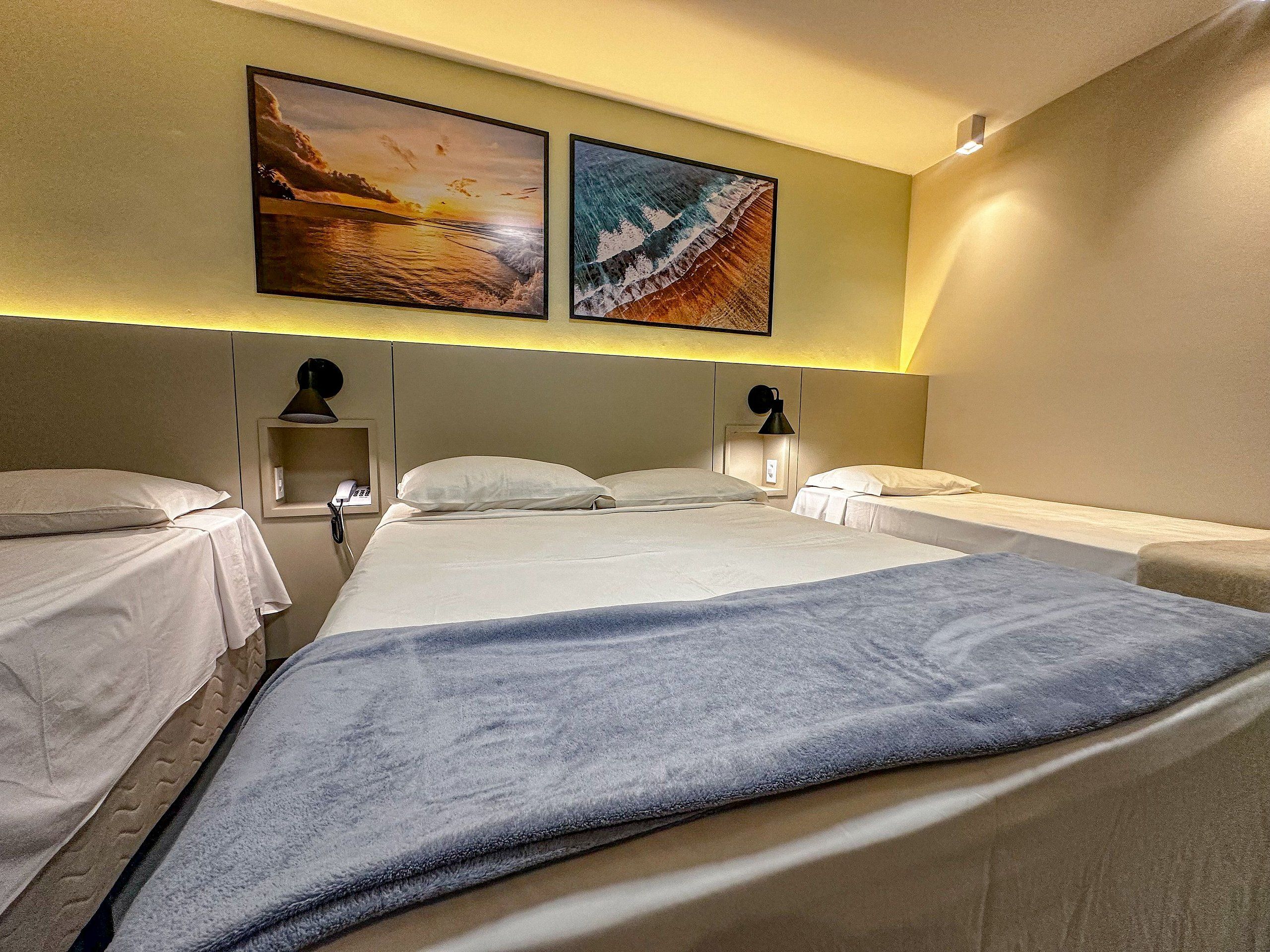 A hotel room with two beds, a large bed with a blue blanket, a smaller bed, a bedside table with a lamp, and two wall-mounted paintings.