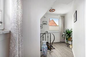 Ein Flur mit einem gemusterten Vorhang, einer Vase, einem Fenster mit Blick auf ein Backsteingebäude und einer Topfpflanze.