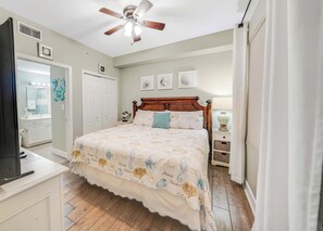 Ein Schlafzimmer mit Bett, Deckenventilator, Nachttisch mit Lampe, Kommode, Spiegel und Tür.