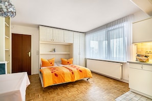 Une chambre avec un plancher en bois, un lit avec une literie orange, une armoire blanche et une fenêtre ornée de rideaux légers.