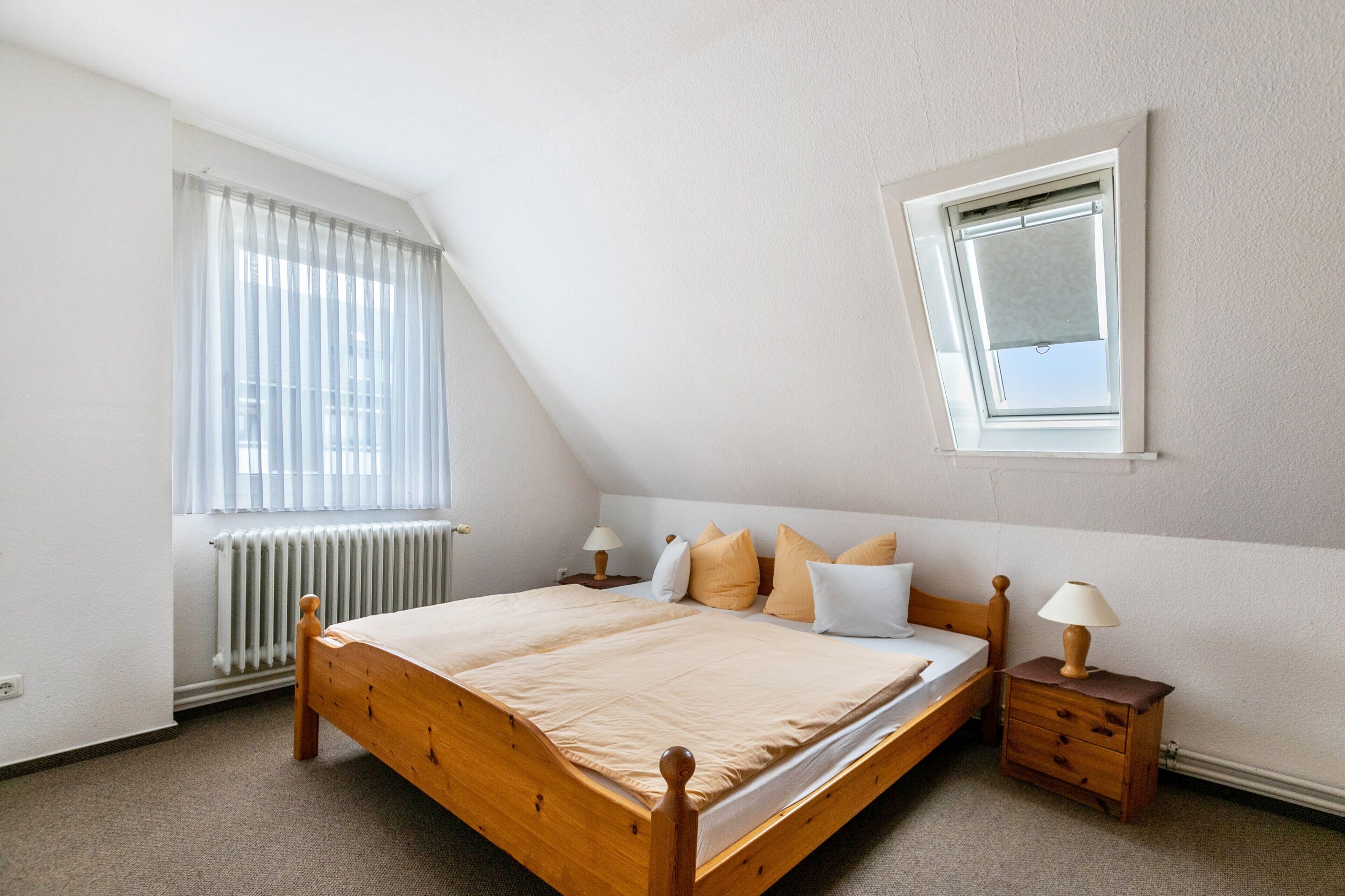 Une chambre avec un plafond en pente, un lit avec un cadre en bois, une table de chevet avec une lampe et une fenêtre avec des stores.
