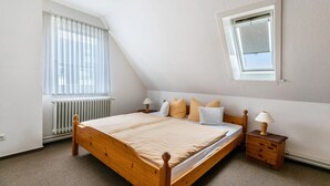 Ein Schlafzimmer mit schrägem Dach, einem Bett mit Holzrahmen, einem Nachttisch mit Lampe und einem Fenster mit Jalousien.