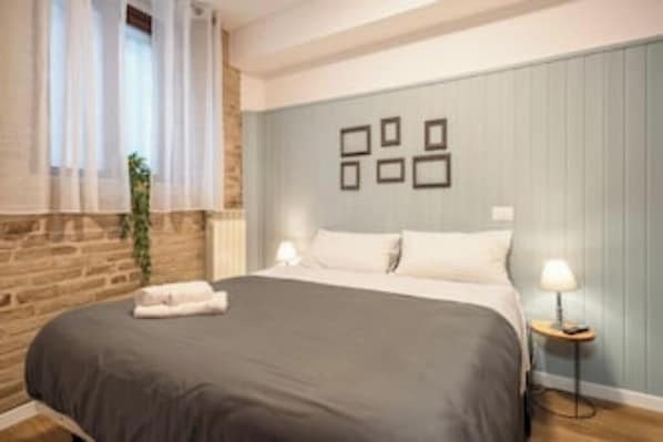 Une chambre à coucher avec un lit, une table de chevet, une lampe fixée au mur et une fenêtre avec des rideaux.