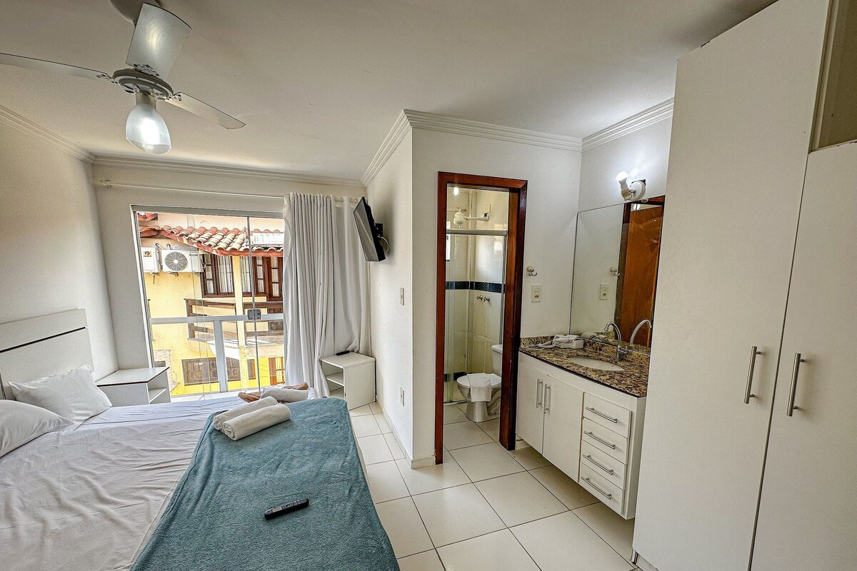 Habitación de hotel con cama, baño con espejo y lavabo, y vistas al balcón exterior.
