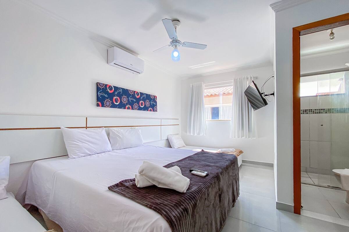 Quarto de hotel com cama, ventoinha no teto, ar condicionado, janela com cortinas e área de chuveiro.