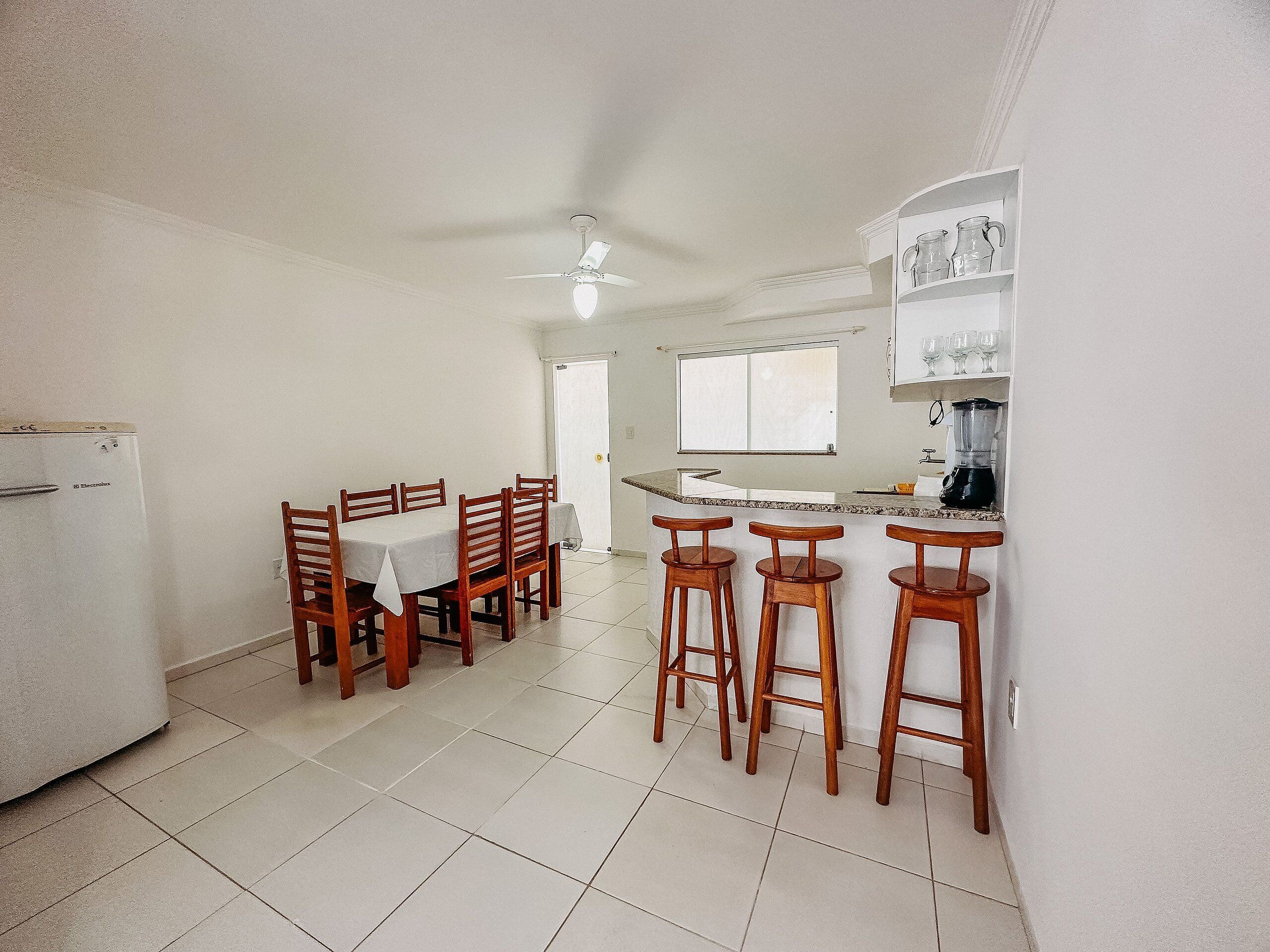 A kitchen with a dining table, bar stools, a refrigerator, a ceiling fan, and a window.