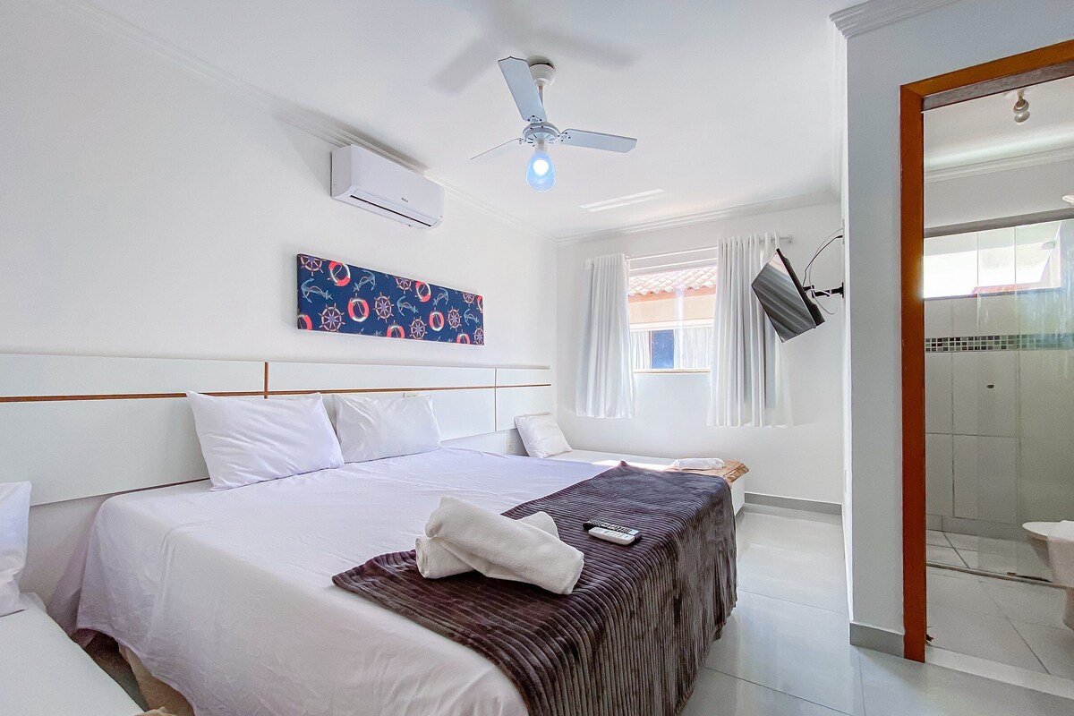 Habitación de hotel con cama, ventilador de techo, equipo de aire acondicionado, ventana con cortinas y zona de ducha.