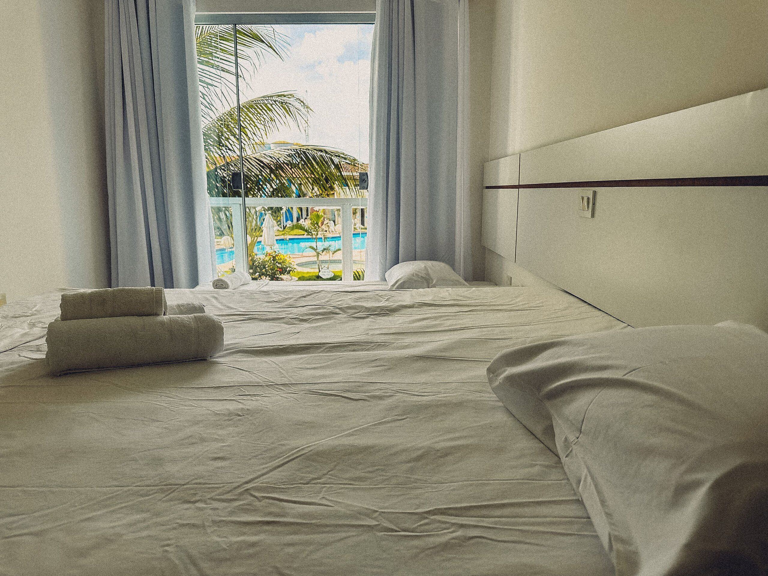 A hotel room with a bed, pillows, and a view of a pool and palm trees.