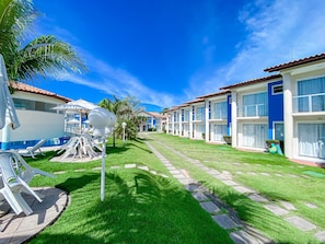 A row of blue and white buildings with a well-maintained lawn, outdoor seating, and a clear sky. - Residencial Mont Sião 1 e 2 (Porto Seguro)