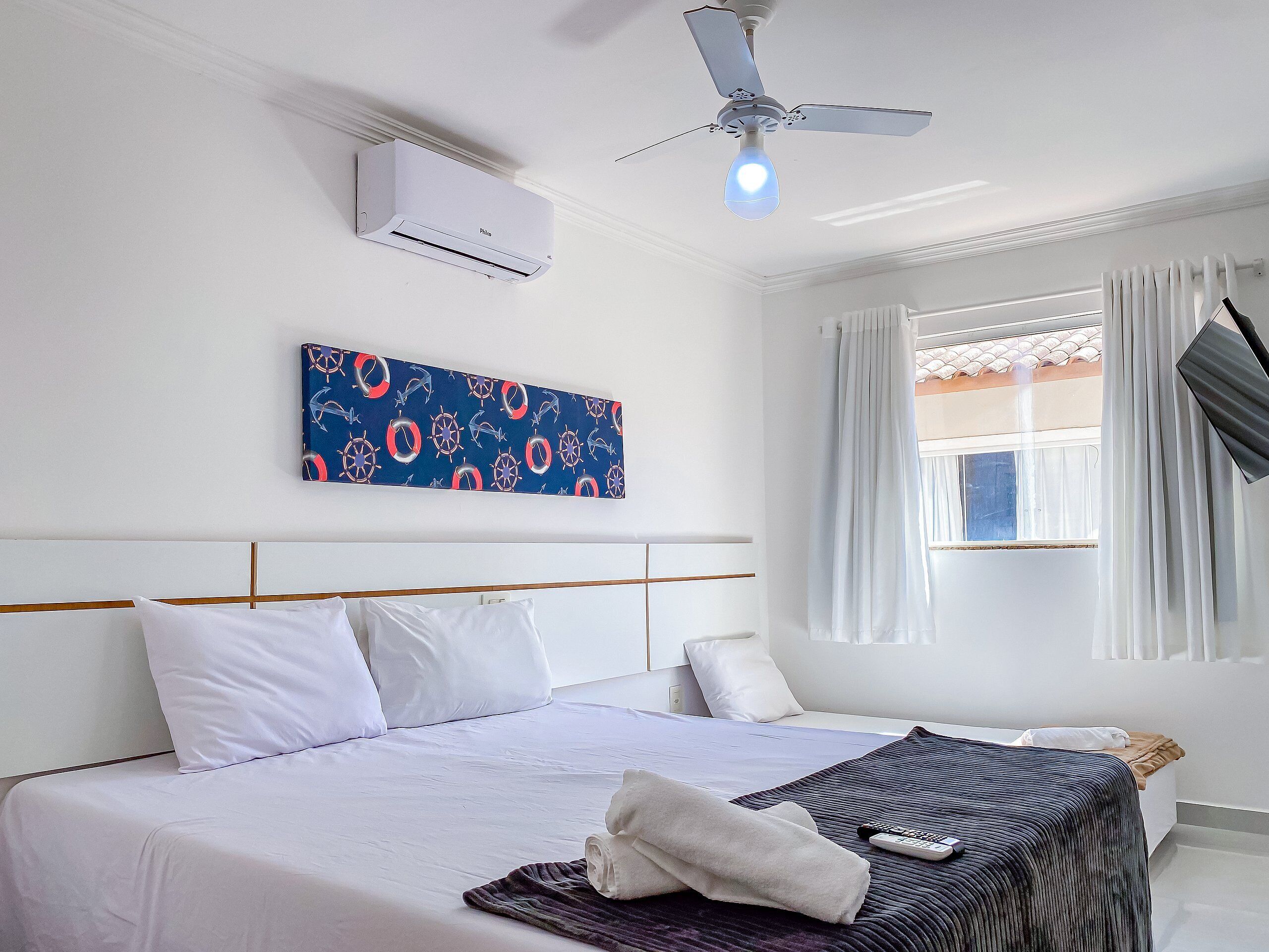 Habitación de hotel con cama, ventilador de techo, equipo de aire acondicionado, ventana con cortinas y una obra de arte colgada en la pared.