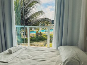 Una habitación con cama, ventana con cortinas y vista a una piscina y palmeras.