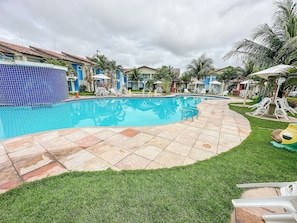 Una piscina con bordes de azulejos, rodeada de sillas de descanso y sombrillas, con césped y edificios al fondo.