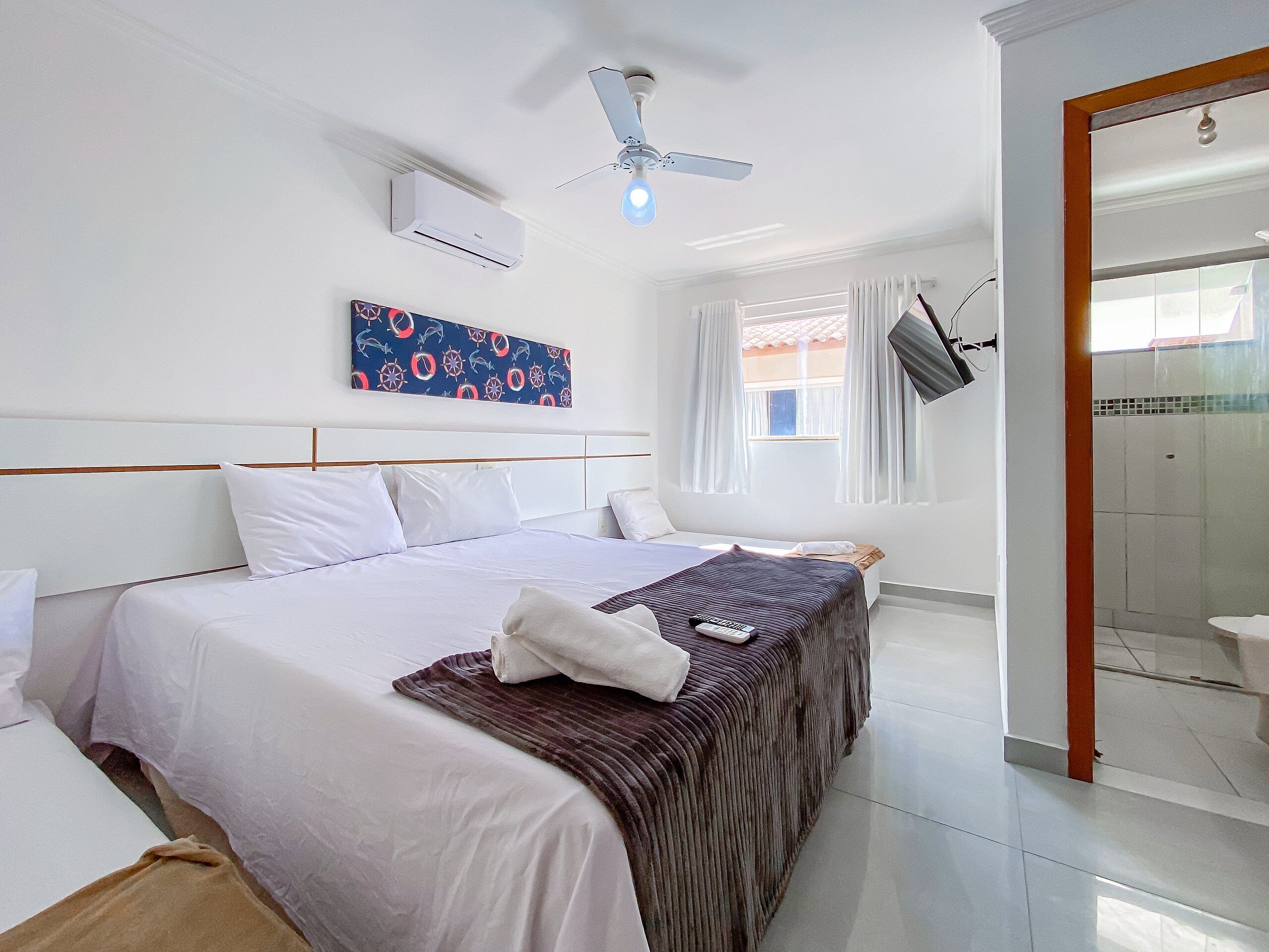 A hotel room with a bed, a ceiling fan, an air conditioning unit, a window with curtains, and a shower area.