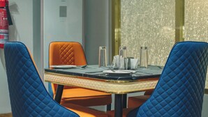 A dining table with two blue quilted chairs, a fire extinguisher, and a window with a textured wall. - Wellington Blu hotel & Suites (Enugu)