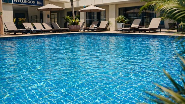 A clear blue pool with lounge chairs and umbrellas, surrounded by a building with a sign reading 'Wellington Blu'. - Wellington Blu hotel & Suites (Enugu)