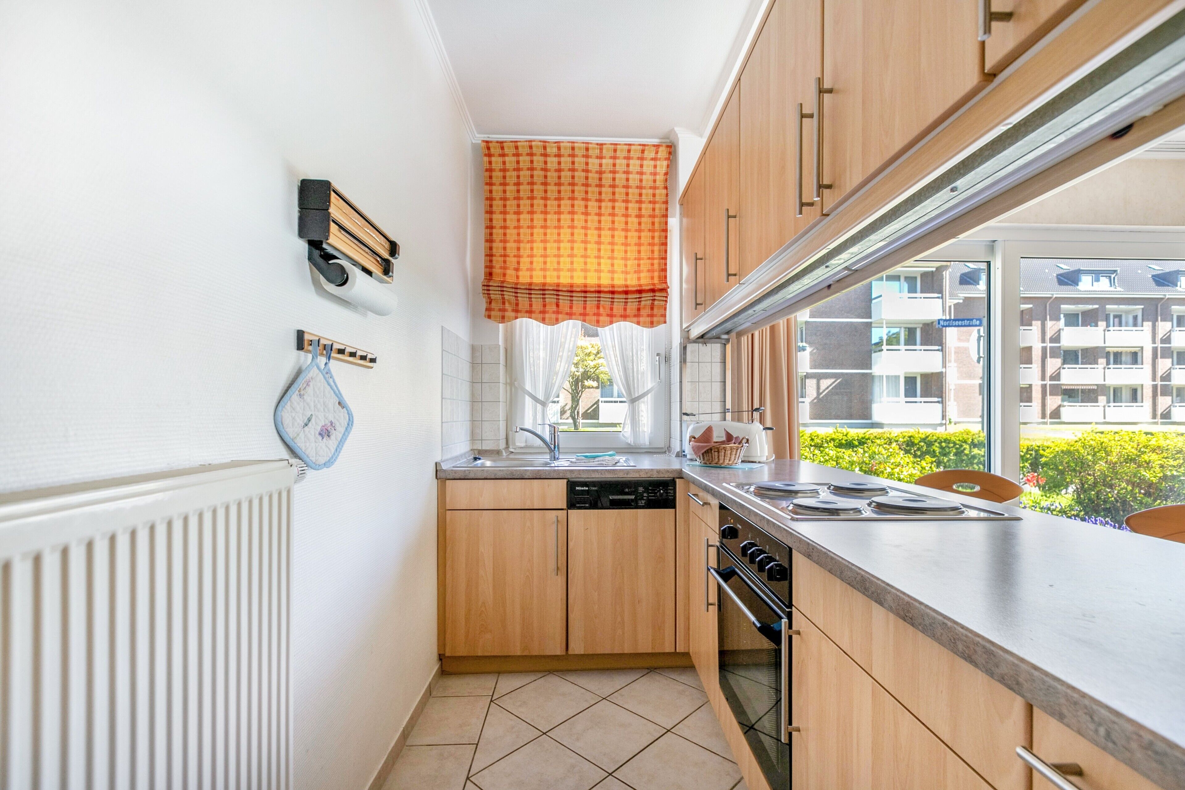 A compact kitchen with wooden cabinets, a dishwasher, a stove, and a window with a checkered blind.