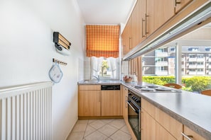 Eine kleine Küche mit Holzschränken, einem Geschirrspüler, einem Herd und einem Fenster mit einem Schachbrettmuster-Jalousie.