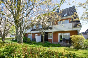 Une maison à deux étages avec un jardin, une terrasse et un petit espace extérieur aménagé pour s’asseoir.