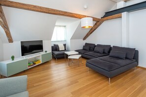 A modern loft-style living room with a large flat-screen TV, a sectional sofa, a small white table, and a wooden ceiling with exposed beams. - Haus Spiegel - Penthouse with roof terrace - Time out (Lindau (Bodensee))