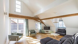 Ein geräumiger Loft mit hohen Decken, freiliegenden Holzbalken, einem großen Fenster und einer modernen Wohnbereich mit Sofa, Fernseher und Sitzgelegenheiten.