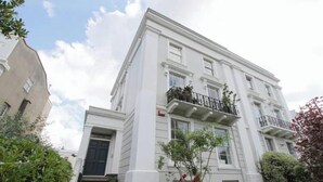 Un edificio blanco de varios pisos con una entrada principal, una escalera y un pequeño árbol en primer plano.