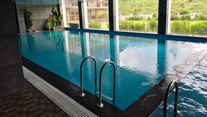 Una piscina interior con paredes de vidrio transparente, suelo de baldosas y vistas a un paisaje verde.