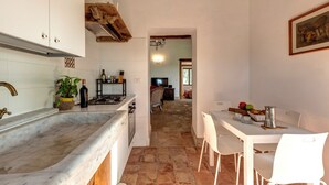 Una cocina con armarios blancos, encimera de mármol, zona de comedor con sillas blancas y un televisor visible al fondo.