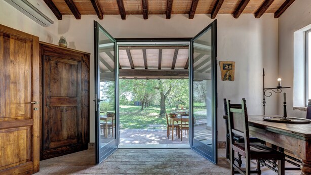 Habitación con puerta de madera, una mesa con sillas, un escritorio y una silla, y vistas a un espacio exterior con árboles y una mesa a través de una puerta de vidrio.