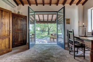 A room with a wooden door, a table with chairs, a desk with a chair, and a view through a glass door to an outdoor area with trees and a table. - Casale il Poderetto by Mamo (Pitigliano)