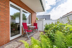 A patio with a brick wall, a sliding glass door, two red chairs, a small round table, and a garden with ferns. - Apartment 'Nr 02' with Wi-Fi (Büsum)