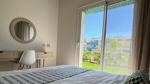 A bedroom with a bed, a desk, a chair, a round mirror, and a large window with a view of buildings and greenery. - Fouka North Coast SA-8A-G03 (Marsa Matrouh)