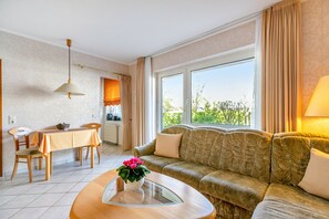 A living room with a sofa, a dining table, chairs, a coffee table with a flower vase, and a window with curtains. - Apartment 'Nr 03' with Wi-Fi (Büsum)